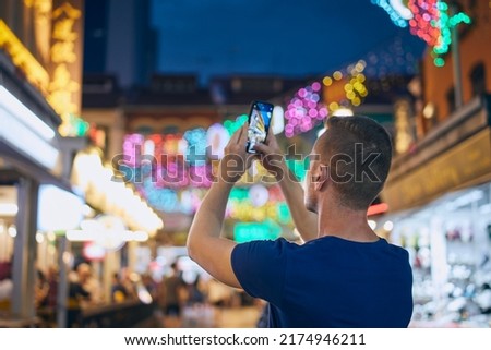 Rear view of man while taking pictures with mobile phone on colorful street. Tourist in Chinetown in Singapore.
