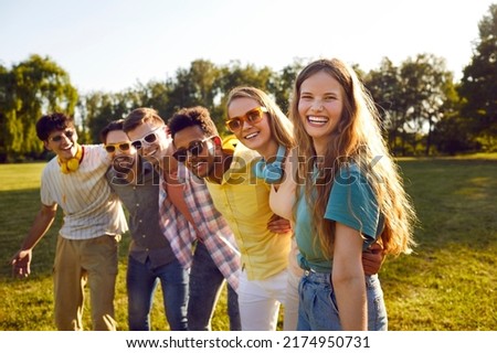 Portrait of happy excited young woman standing with her multiracial friends in green park. Joyful woman standing in row with cheerful male and female friends who are wearing sunglasses. Youth concept. Royalty-Free Stock Photo #2174950731