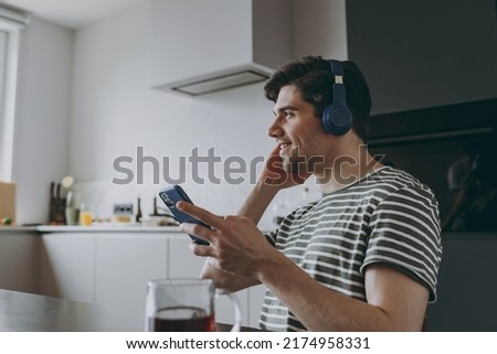 Young side view smiling happy man in striped t-shirt headphones using mobile cell phone look aside listen to music browsing sitting by table in light kitchen at home alone. People lifestyle concept.