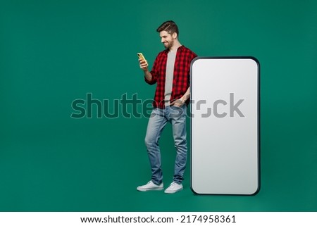 Full body young smiling man he 20s wearing red shirt grey t-shirt big huge blank screen mobile cell phone with workspace copy space mockup area use smartphone isolated on plain dark green background.
