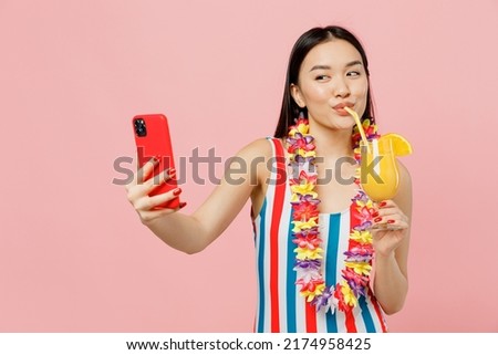 Young woman of Asian ethnicity in striped one-piece swimsuit hawaii lei hold cocktail do selfie shot on mobile cell phone isolated on plain pastel pink background. Summer vacation sea rest concept.