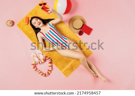 Top view full body young woman of Asian ethnicity in striped swimsuit lies on towel hotel pool do selfie shot mobile cell phone isolated on plain pastel pink background. Summer vacation sea concept