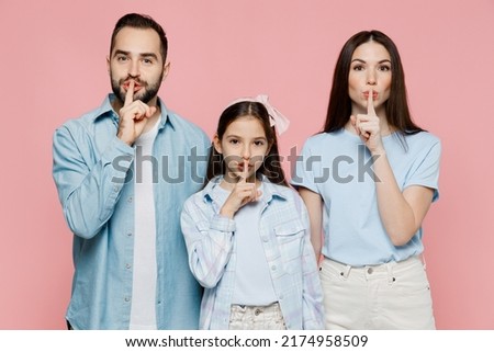 Young parents mom dad with child kid daughter teen girl in blue clothes say hush be quiet with finger on lips shhh gesture isolated on plain pastel light pink background. Family day parenthood concept