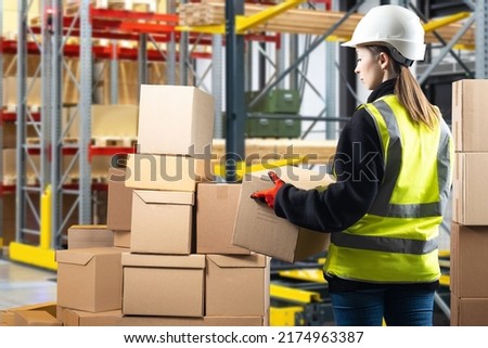 Warehouse manager. Woman with box inside logistics warehouse. Warehouse company manager career concept. Distribution center manager in yellow vest. Girl in hardhat in front of blurred shelving
