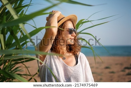 Portrait of a young cheerful woman in a hat sitting on the seashore at sunset with a smartphone in her hands