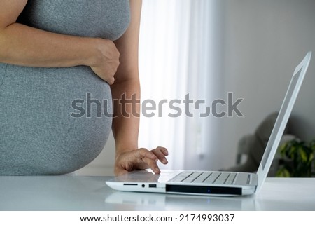 Pregnant woman with big belly use her white laptop on table at home interior. Browsing, surfing net, do online shopping, reading maternity blogs or chatting online. Pregnancy and technology.