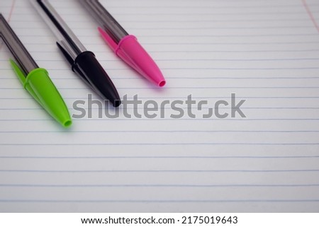 Close-up of three pens with caps on resting on a lined paper sheet. Background of green, black and pink pens with selective focus and empty space for text Royalty-Free Stock Photo #2175019643