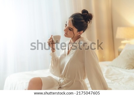 Calm brunette woman in white silk bathrobe sitting on bed and drinking coffee at the sunny morning bedroom. Me time concept Royalty-Free Stock Photo #2175047605