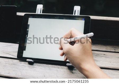 Young creative designer holding stylus pen drawing on screen of digital tablet while sitting at the park.