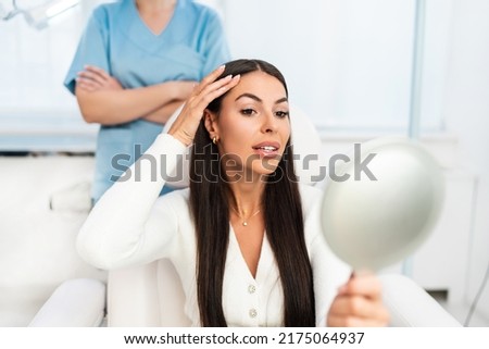 Beautiful and happy young woman sitting in medical chair and looking in the mirror. She is satisfied after successful beauty treatment with hyaluronic acid fillers or botulinum toxin injections. Royalty-Free Stock Photo #2175064937