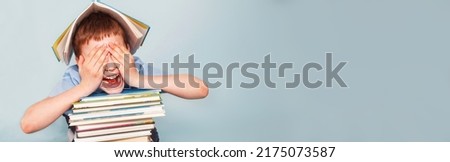 Upset schoolboy sitting with pile of school books and covers his face with hands isolated on a blue background