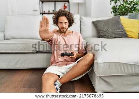 Young hispanic man playing video game holding controller at home with open hand doing stop sign with serious and confident expression, defense gesture 