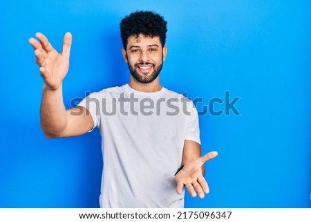 Young arab man with beard wearing casual white t shirt smiling cheerful offering hands giving assistance and acceptance. 