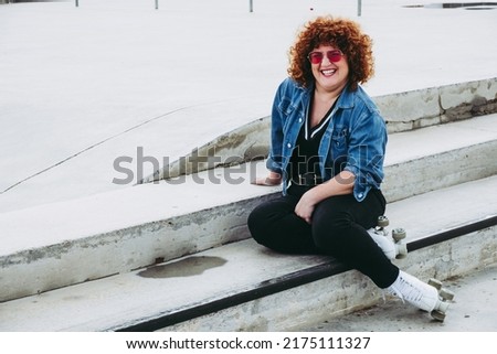 Curvy woman on roller skates does sports outdoors. Body positive chubby lady is exercising in the park. Smiling and happy young lady wears vintage denim clothes and colored glasses.