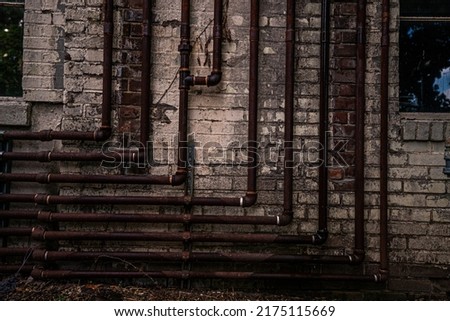 Rusty metal pipes forming a backwards L shape attached to w weathered white brick wall.
