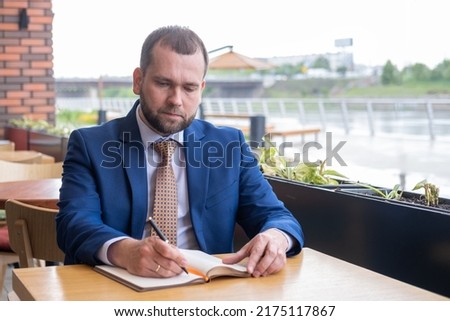 Focused middle aged 40s businessman writes notes in a personal diary, plans a working day, checks a schedule, or writes important information by hand in a street cafe. Business meeting planning