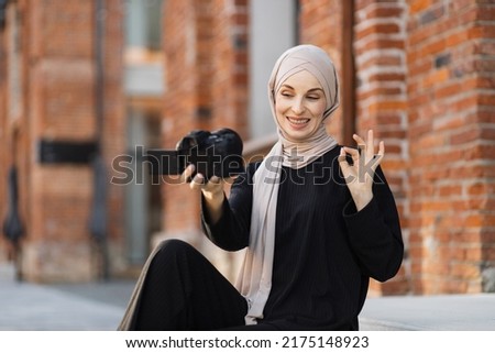 Young muslim woman using a video camera to record video for her work. Happy and excited arabic business woman wear black clothes with hijab, waving to internet audience. Influencer vlogging concept