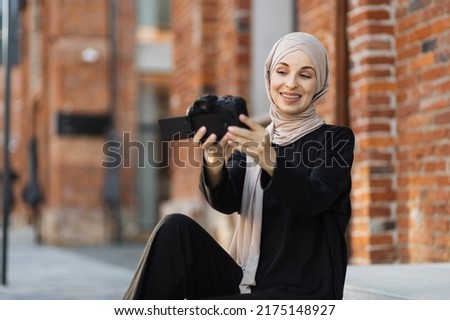 Young female muslim blogger in trendy clothes and hijab sitting on stairson city street, records herself on video holding camera in hand. Arab girl takes pictures of herself on camera.