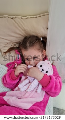An emotional little girl lies on a light sofa in a pink plush pajama suit and hugs a pink bunny toy. The child goes to bed and falls asleep with a toy in his hands. Mom is reading a story.