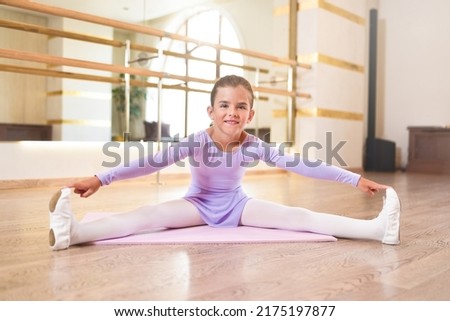 Beautiful little ballerina in a lilac leotard and white tights doing stretching exercises on a pink karemat in dance studio.