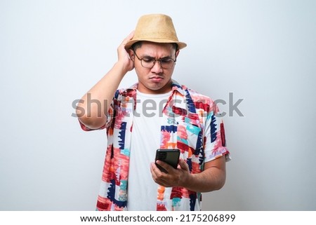Portrait of unhappy Asian man in casual beach shirt holding mobile phone with sad expression on face while scratching his head. Isolated image on white background Royalty-Free Stock Photo #2175206899