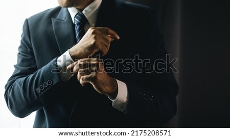 Confident businessman buttoning or adjust classic blue suit near window in hotel room at the morning. Handsome man wearing a nice suit on wedding day. Royalty-Free Stock Photo #2175208571