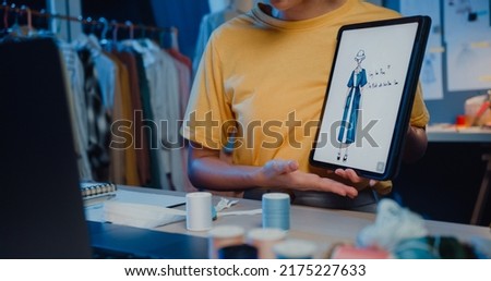 Close-up young Asia lady dressmaker use digital tablet meeting with designer discuss show sample dress design new collection on screen on desk in shop at night. Tailor new business startup concept.