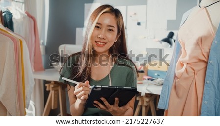 Cheerful professional Asia lady fashion designer with casual use digital tablet create new collection in her shop. Lady tailor study draw sketch on tablet pad smile look at camera in workshop studio.