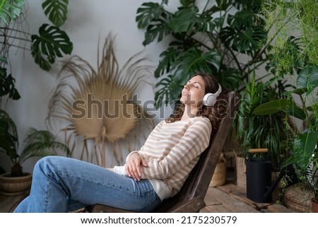 Beautiful young woman in wireless headphones sits with close eyes on wooden chair in urban jungle garden relax space. Calm female enjoying good meditative music, online radio, audiobook or podcasts.