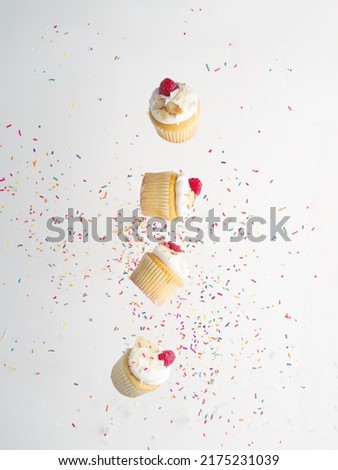 Small mouth-watering muffins with cream and raspberries on a white background with multi-colored confetti in a frozen flight. Festive composition. Banner, advertising. Royalty-Free Stock Photo #2175231039
