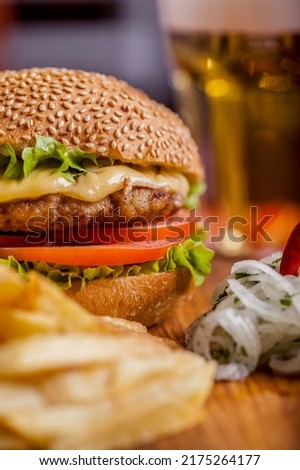 Burger with sesame bun with onions, salad, meat cutlet, tomatoes and cheese on a wooden board with a glass of light beer. Vertical orientation