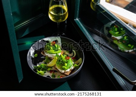 Sandwiches with avocado, beans, chukoy, philadelphia cheese, parsley, pepper and white sauce in a black plate against the background of a dark green window with a glass of wine