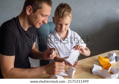 A cute boy is sitting with his dad at the table and collecting origami. Glue the parts