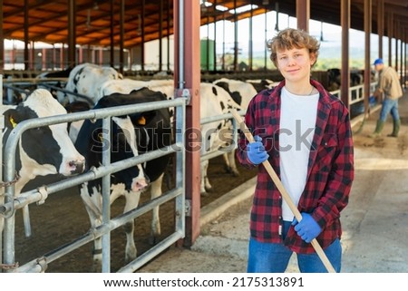 Young male milk farm worker standing in cowhouse with working tool in hands. Royalty-Free Stock Photo #2175313891