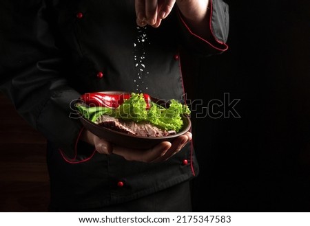 The chef sprinkles salt on the sliced meat with vegetables on the plate. Food preparation concept on black background