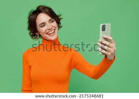Young smiling happy woman 20s wear casual orange turtleneck doing selfie shot on mobile cell phone post photo on social network isolated on plain pastel light green color background studio portrait.