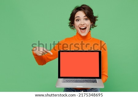 Young smiling happy woman 20s wear orange turtleneck hold use work point finger on laptop pc computer with blank screen workspace area isolated on plain pastel light green background studio portrait.