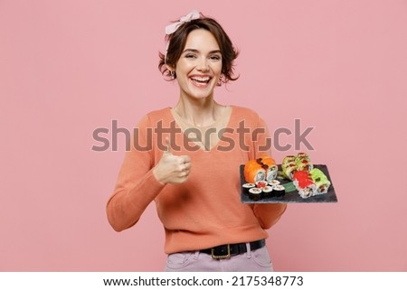 Young satisfied woman 20s in casual clothes hold in hand makizushi sushi roll served on black plate traditional japanese food showing thumb up like gesture isolated on plain pastel pink background.