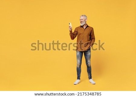 Full size body length excited confident smiling elderly gray-haired bearded man 50s years old wears brown shirt hold in hand use mobile cell phone isolated on plain yellow background studio portrait