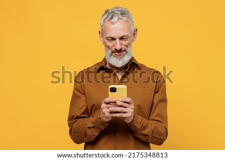 Happy excited elderly gray-haired bearded man 50s years old he wears brown shirt hold in hand use mobile cell phone texting typing searching reading isolated on plain yellow background studio portrait