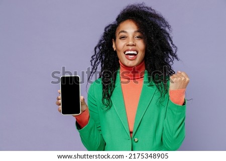 Young black curly woman 20s wears green shirt hold in hand use mobile cell phone with blank screen workspace area doing winner gesture isolated on plain pastel light violet background studio portrait