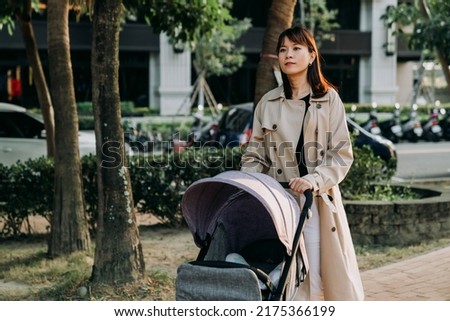 portrait happy chinese career mom is pushing walking with a baby pram at leisure in a park at urban area on an autumn sunny day.