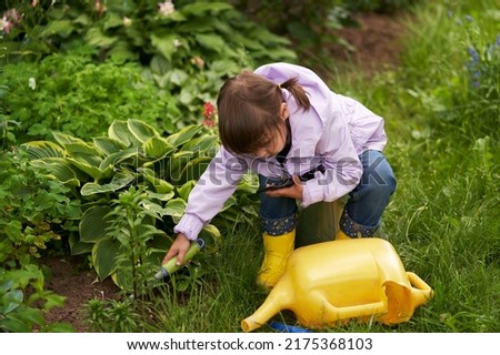 The girl is raking the ground on a flower bed in the garden with one hand, holding a smartphone in the other hand and watching a cartoon.                               