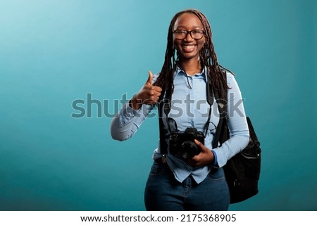 Cheerful professional photography entusiast with DSLR device saying is ready for photo shooting by giving thumbs up sign. Joyful photographer giving ok finger gesture while standing on blue background