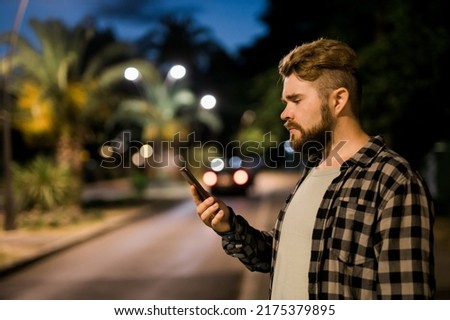 Bearded man holding scrolling texting in his cellphone at night street. Guy calls for taxi in an app in evening city - copy space and place for advertising