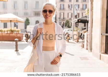 Charming european woman smilies at camera wearing white costume . Caucasian blonde in sunglasses on the background of city 