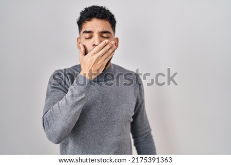 Hispanic man with beard standing over white background bored yawning tired covering mouth with hand. restless and sleepiness. 