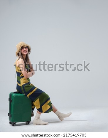Happy Asian woman traveler siting and holding suitcase isolated on gray background, Tourist girl having cheerful holiday trip concept, Full body composition