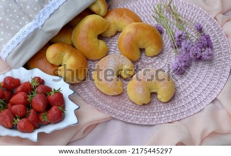 Home baked sweet buns, small breads with strawberries and violet flowers on light pink tablecloth. Delicious sandwich breads and fruit bowl. Fresh baked bread rolls with soft crust, fluffy crumb.