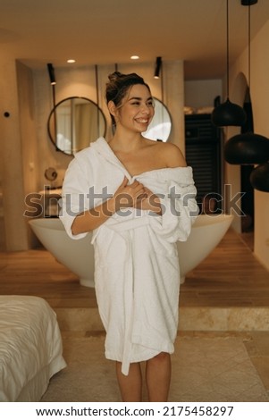 Cheerful blonde girl wearing bathrobe rejoicing at her cozy bathroom after beauty routine. Me time concept. Stock photo Royalty-Free Stock Photo #2175458297